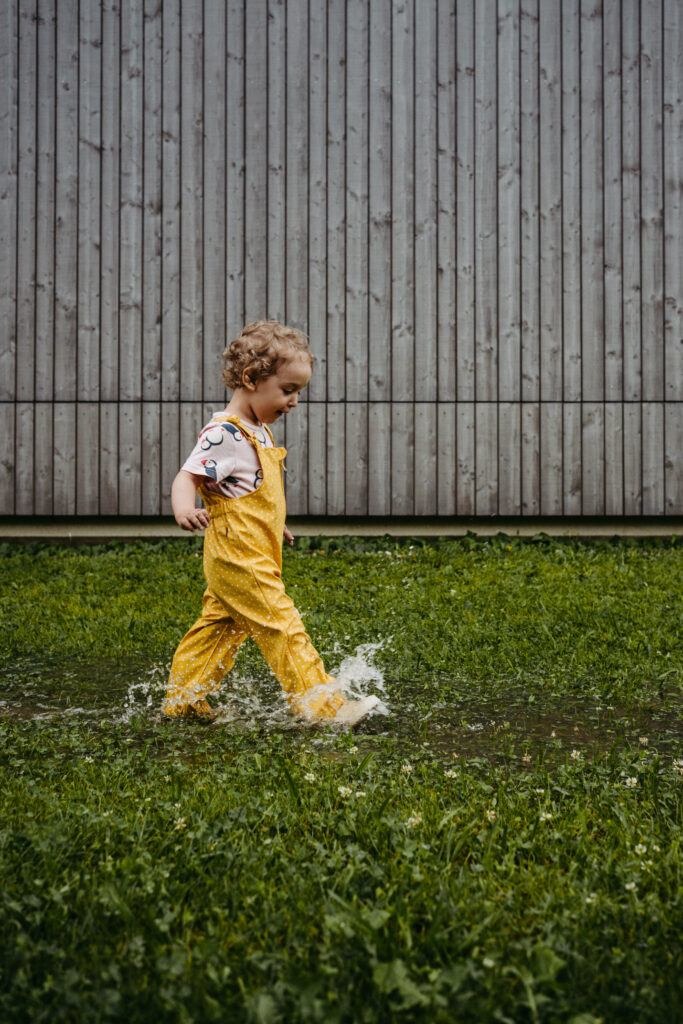 Mädchen in gelber Regenhose läuft durch eine Lacke – natürliche Familienfotos Graz und Umgebung, authentische Familienfotografie