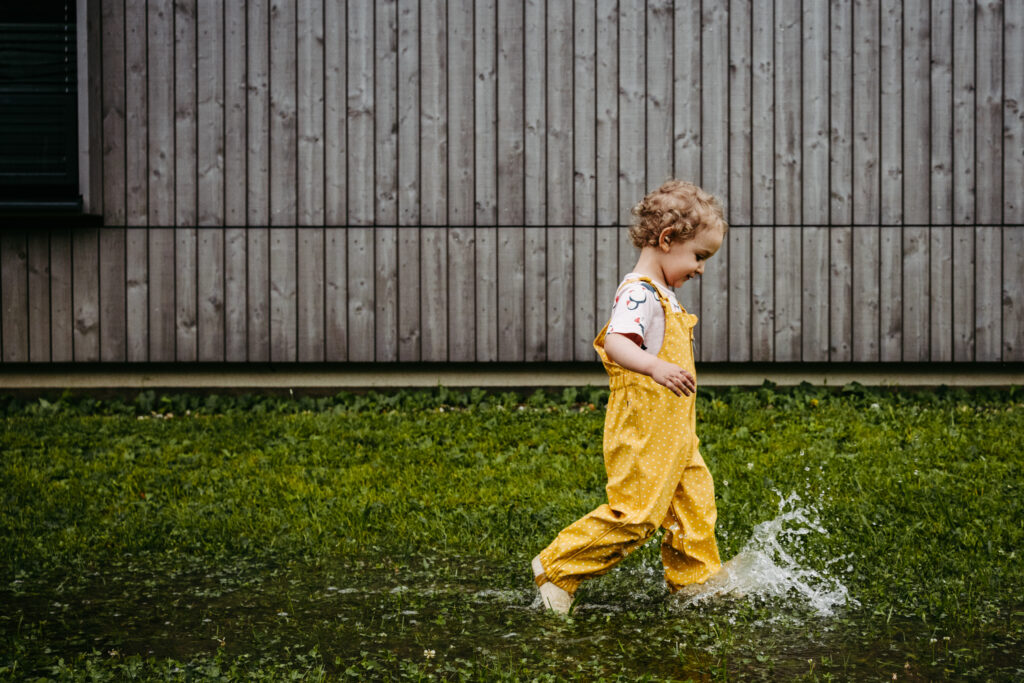 Mädchen in gelber Regenhose läuft durch eine Lacke – natürliche Familienfotos Graz und Umgebung, authentische Familienfotografie