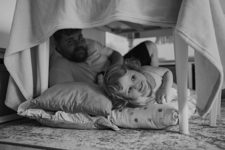 Mädchen liegt mit Papa unter dem Küchentisch und lacht hervor – dokumentarische Familienfotos Graz und Umgebung