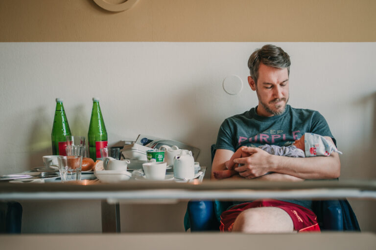Papa hält neugeborenes im arm mit chaotischem Frühstückstisch im Hintergrund. dokumentarische Familienfotografie graz