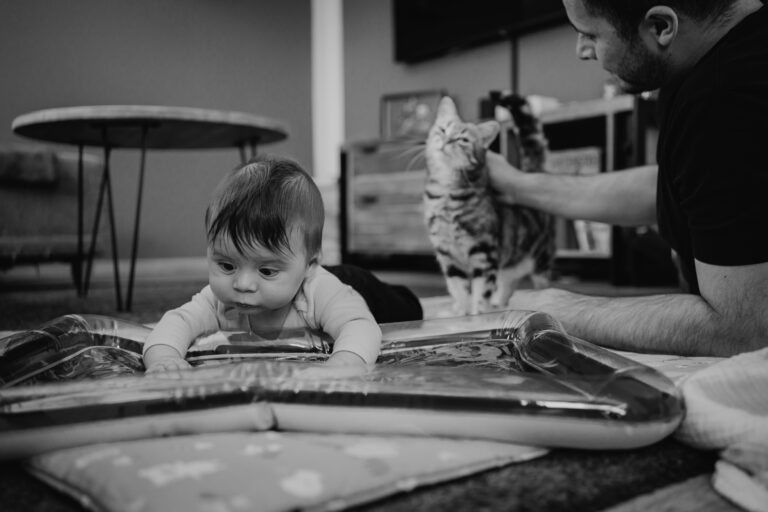 Baby übt Bauchlage, Papa streichelt die Katze im Hintergrund – natürliche Familienfotos Graz und Umgebung, authentische Alltagsszenen