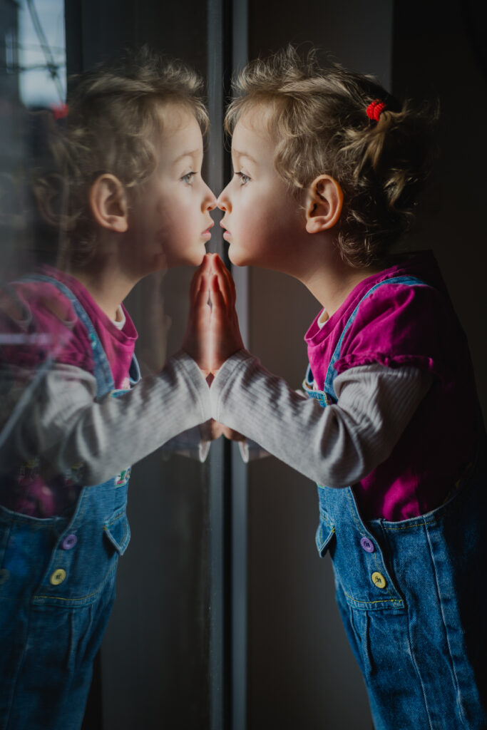 Mädchen blickt in die Fensterscheibe und spiegelt sich darin – dokumentarische Familienfotografie in Graz und Graz Umgebung