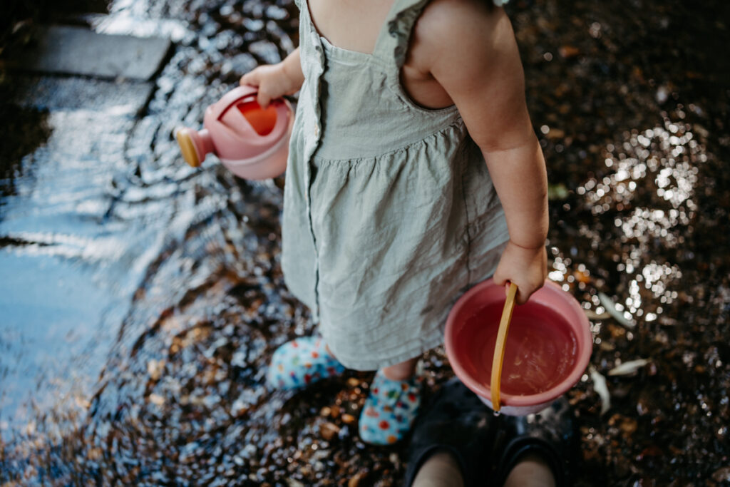 Oberansicht eines Mädchens, das mit Kübel und Gießkanne im Bach steht – spielerischer Alltagsmoment, dokumentarische Familienfotografie in Graz und Umgebung