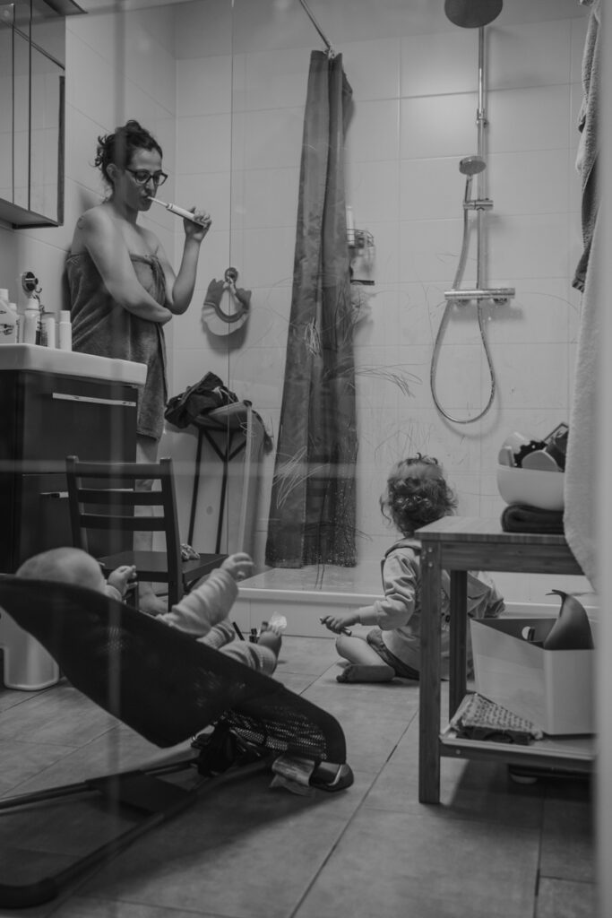 Mama steht Zähneputzend im Bad nur mit Handtuch, während ihre Kinder spielen. Echte Familienfotografie Graz und Umgebung.