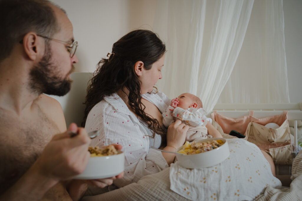 Eltern liegen mit Neugeborenem im Bett und sind selbst gerade beim Frühstücken. Echte Familiengeschichten fotografiert in Graz.