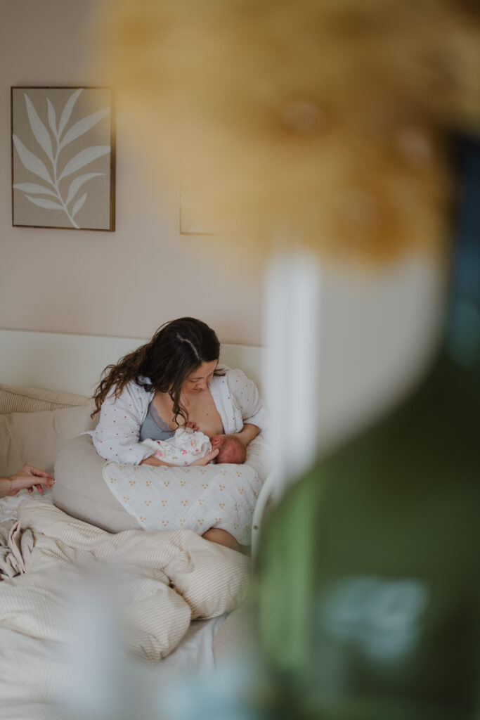 Mama sitzt im Bett und stillt ihr Neugeborenes. Authentische Familienfotografie aus dem Wochenbett in Graz.