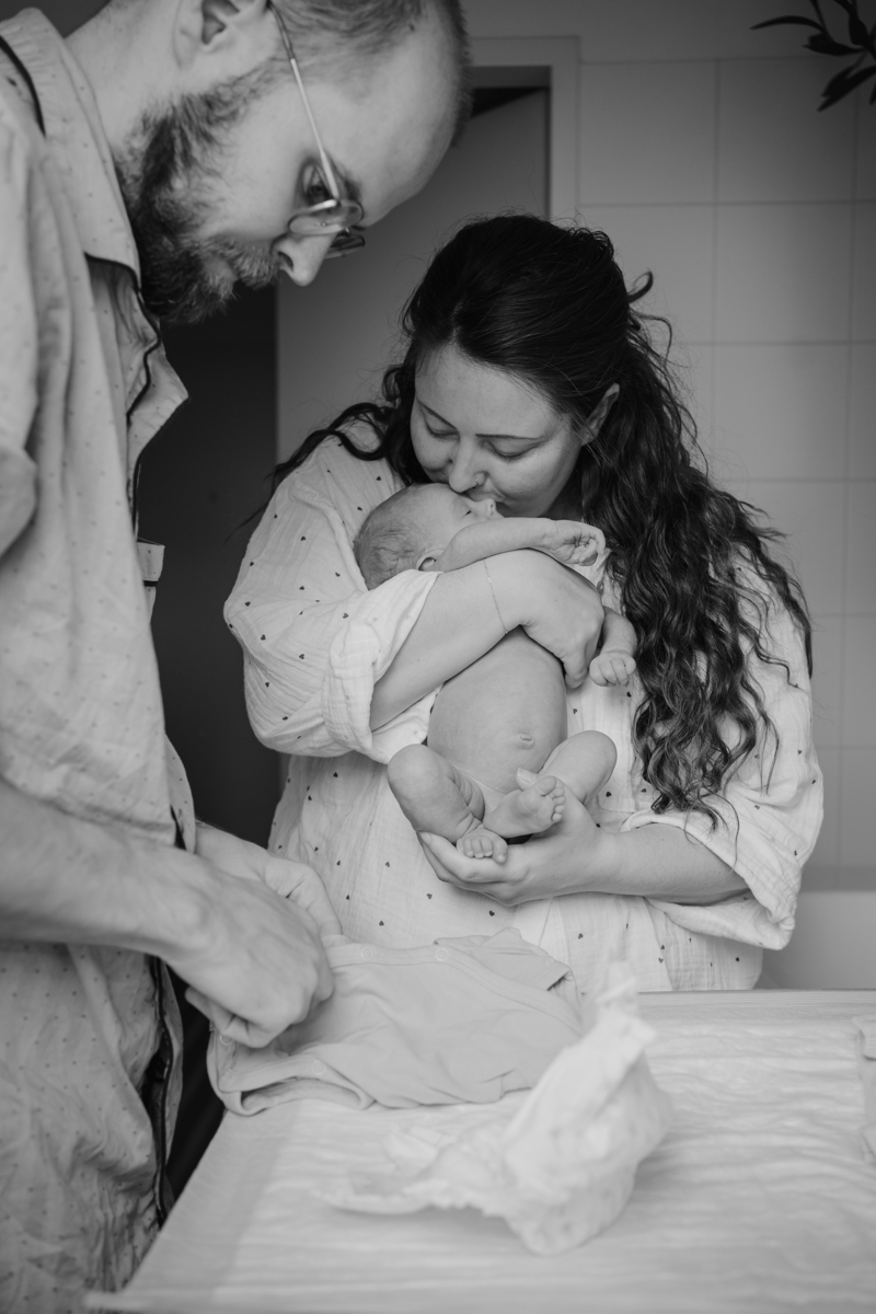 Eltern stehen im Badezimmer, Mama küsst Baby auf den Kopf. Echte Erinnerungen aus einem Wochenbett Fotoshooting in Graz.