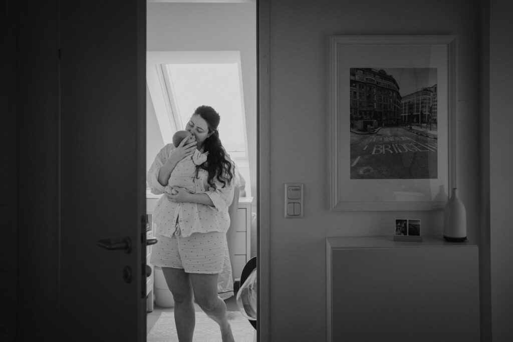 Mama steht mit Neugeborenem im Badezimmer. Natürliche Aufnahme während Wochenbett Fotoshooting in Graz.