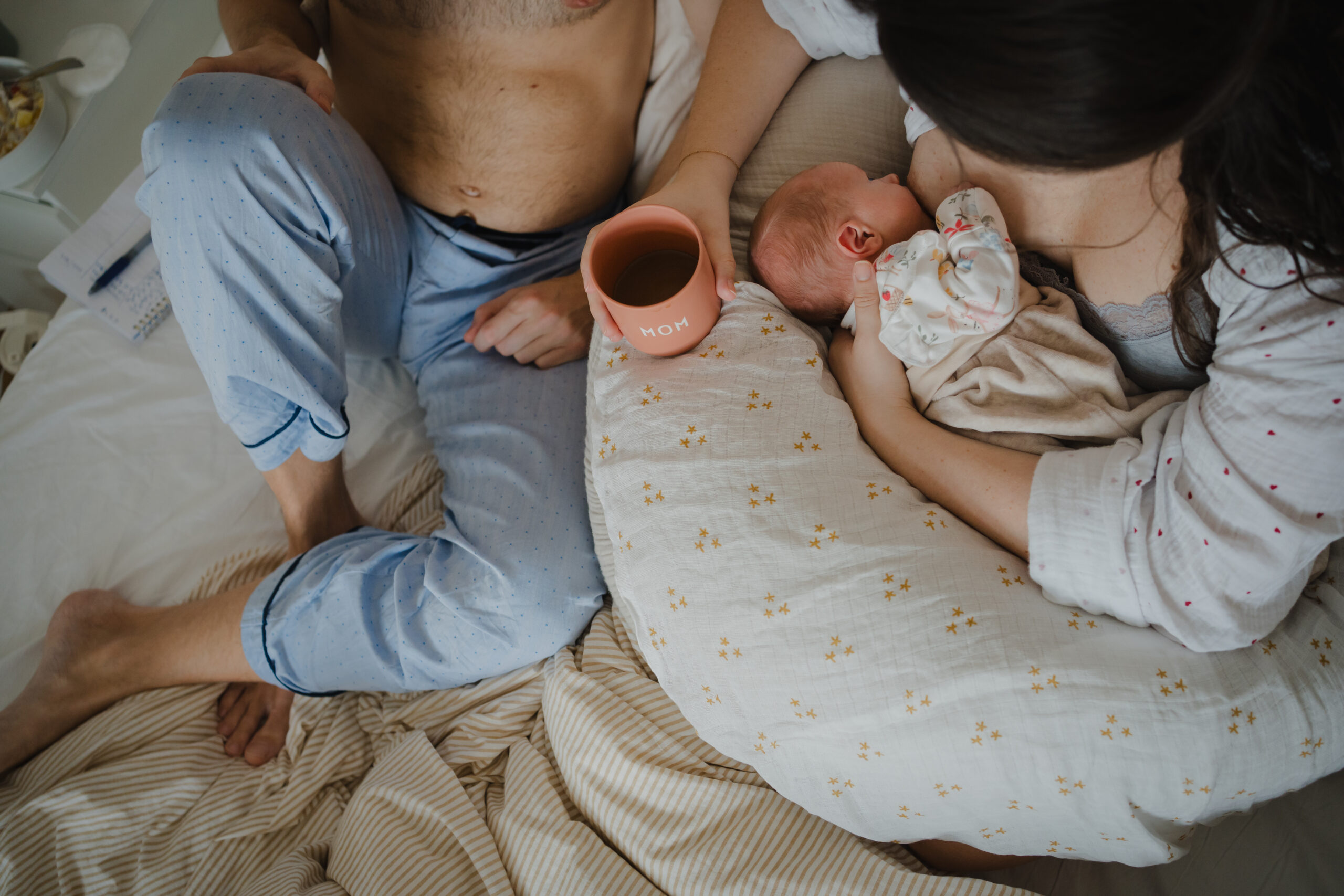 Obenansicht von einer Familie die mit Neugeborenem im Bett frühstückt. Authentische Familienfotografie während dem Wochenbett.