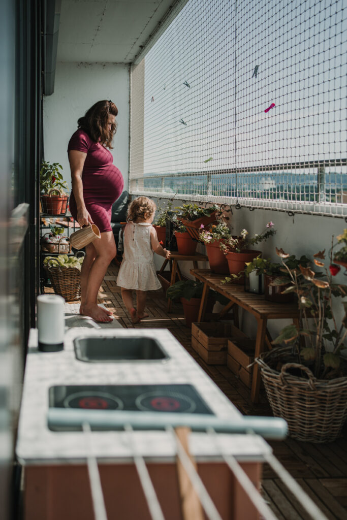 eine schwangere Mama ist mit ihrem Kleinkind am Balkon und gießt die Blumen, so sieht authentische Schwangerschaftsfotografie in Graz und Umgebung aus.