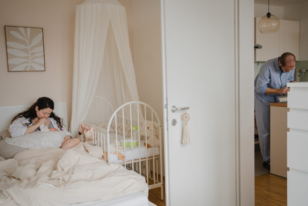 Mama liegt mit Neugeboren im Bett gleichzeitig sieht man den Papa in der Küche das Frühstück vorbereiten. dokumentarische Familienfotografie in Graz.