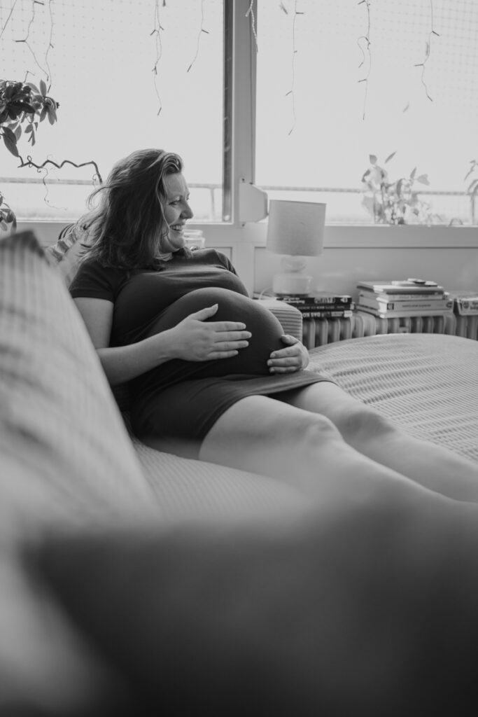 Schwangere Mama liegt am Sofa und hält ihren Brauch während sie lacht. Natürliche Schwangerschaftsfotografie Graz.