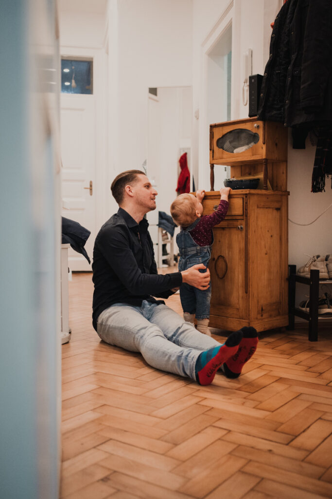 Papa hält Kind fest, während es versucht auf ein Regal zu klettern. Authentische Familienfotografie Graz.