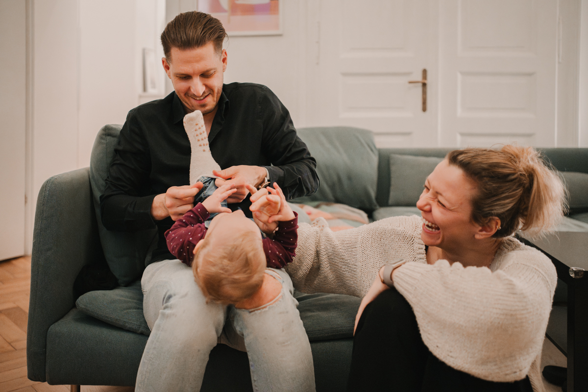 Papa hat Kind am Schoß und versucht ihm die Socken anzuziehen, Mama sitzt daneben und lacht. Angestellte Familienfotografie in Graz und Umgebung.