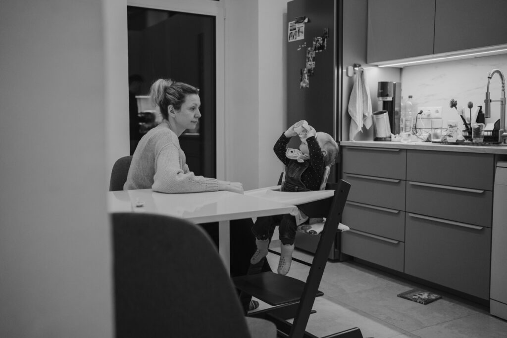 Mama sitzt mit Kind am Küchentisch und beobachte es, wie es aus Flasche trinkt. Ungestellte Familienfotografie in Graz und Umgebung.