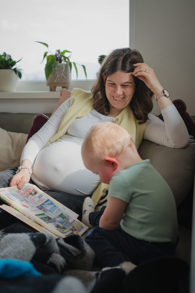 Schwangere Mama sitzt mit Kleinkind am Sofa und liest eine Zeitschrift. Authentische Babybauchfotografie in Graz.