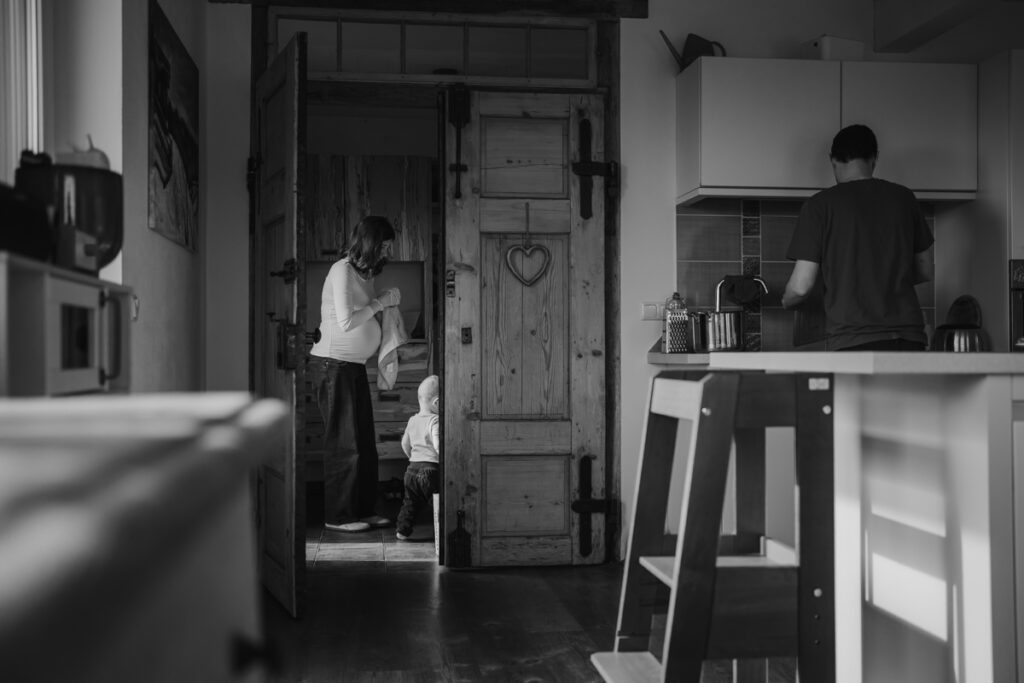 Schwangere Mama steht mit Kleinkind im Vorzimmer, während Papa in der Küche kocht. Schwangerschaftsfotografie in Graz.