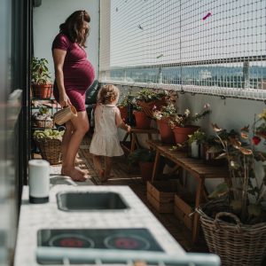 eine schwangere Mama ist mit ihrem Kleinkind am Balkon und gießt die Blumen, so sieht authentische Schwangerschaftsfotografie in Graz und Umgebung aus.